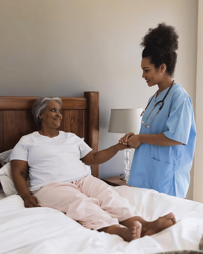 A hospice care nurse holding hands with an elderly patient on a bed.